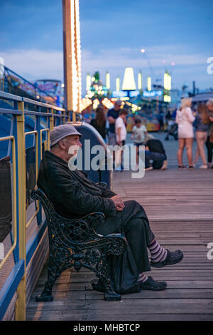 Un eldelry figure masculine assise sur un banc sur la jetée de Brigton touchant son pied pour la musique forte alors que les jeunes gens crier sur les manèges Banque D'Images