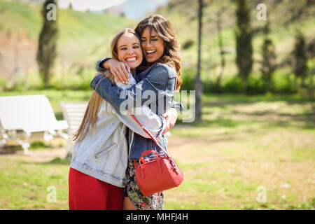 Deux filles heureux hugging in parc urbain. Blonde et brunette filles portant des vêtements à l'extérieur. Banque D'Images