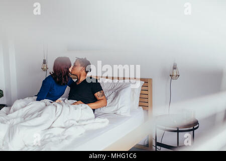Couple romantique au lit. L'Interracial l'homme et la femme assis sur le lit et s'embrassent. Banque D'Images