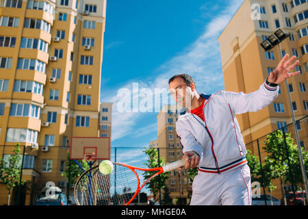 Déterminé beau mâle joueur de tennis jouer au tennis, de frapper la balle, le court de tennis ensoleillé, à l'arrière-plan de la ville Banque D'Images