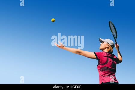 Fond voir le jeune femme, joueur de tennis jouer au tennis, au service de la balle sur bleu ciel avec l'espace vide. Banque D'Images