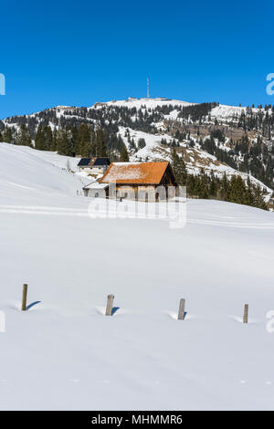 Vieille maison de bois debout dans la neige profonde sur une pente de montagne Rigi avec vue sur pic Rigi-Kulm avec d'énormes antennes télécom sur une bonne journée de printemps Banque D'Images