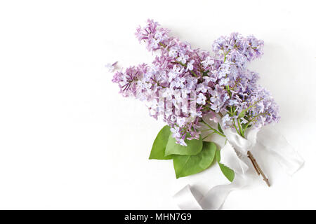 Stock photo style. Printemps mariage, anniversaire scène, composition florale. Bouquet de fleurs fait belle lilas pourpre branches, ruban de soie isolé sur fond de table blanc. Mise à plat, vue du dessus Banque D'Images