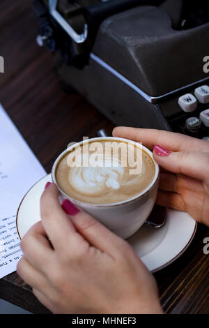 Concept d'écriture. Femme mains tenant une tasse de cappuccino sur un bureau avec une machine à écrire rétro et du papier. Composition verticale. Banque D'Images