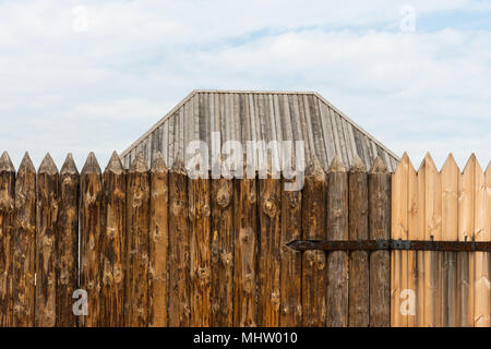Les journaux ont souligné d'une ancienne clôture pâle ou palissade. Un toit en bois d'un bâtiment derrière la clôture. Charnière de porte en métal rouillé. Ciel nuageux Banque D'Images