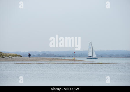 Un jeune couple avec leur bébé à la broche ou la binette à West Wittering avec un yacht de voile en arrière-plan sur une journée sans nuages avec l'ouest vers le bas Banque D'Images