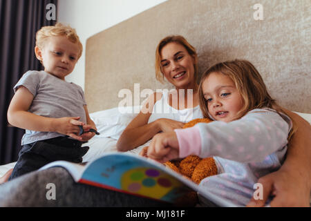 Jeune maman avec ses enfants sur le lit lisant un livre d'histoires. Cute little girl pointing à storybook alors qu'il était assis avec sa mère et son frère sur le lit. Banque D'Images