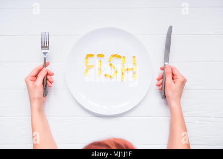 Vue d'en haut les mains tenant le couteau et la fourchette sous plaque ronde avec lettrage mot de poisson par l'huile de foie de morue Capsules Omega 3, sur la table en bois blanc. Banque D'Images