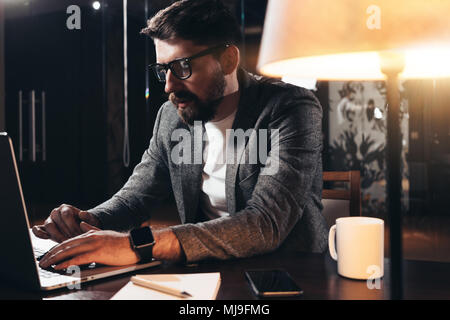 Jeune homme barbu travaillant dans l'espace loft dans la nuit. Un collègue est assis par la table en bois avec lampe et outils office. Directeur de projet à l'aide d'contempo Banque D'Images