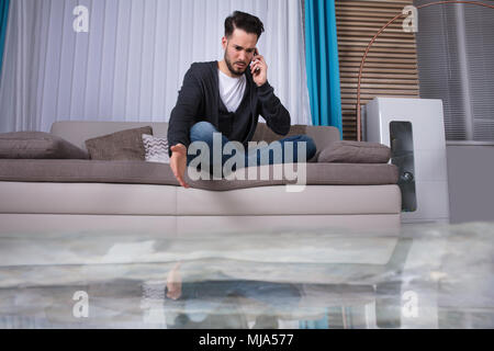 Sol inondé de fuites d'eau en face de l'homme assis sur le canapé à appeler plombier Banque D'Images