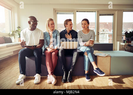 Groupe diversifié de businesspeople smiling et parler ensemble en position assise sur une ligne dans un bureau moderne et lumineux Banque D'Images