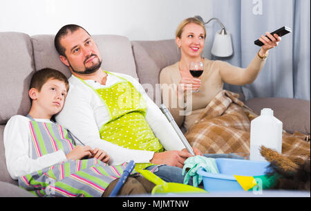 Père fatigué avec fils habillé pour le nettoyage assis sur un canapé, tandis que mère regarder la télévision Banque D'Images