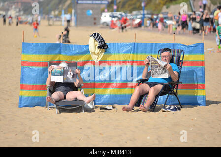 Boscombe, Bournemouth, Dorset, Royaume-Uni, 6th mai 2018 : couple assis sur des chaises avec leurs pieds en lisant les journaux et se détendant sur la plage sous le soleil chaud protégé par une brise-vent. Le week-end des fêtes le plus chaud du Mayday en date. Banque D'Images