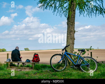 Winton Estate, East Lothian, Ecosse, Royaume-Uni, 6 mai 2018. Laisser les cyclistes de vélos s'appuyant sur les arbres, tandis qu'un homme plus âgé est assis sur un banc de la cycliste et jouit de la vue sur la campagne à travers un champ labouré Banque D'Images