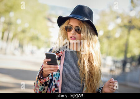 Young woman looking at her smartphone en contexte urbain. Girl wearing hat, veste, pull et lunettes de soleil. Banque D'Images