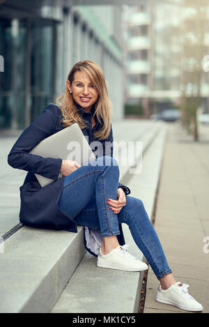 Trendy attractive blonde woman en jeans assis sur les marches extérieures de la ville tenant un ordinateur portable à l'appareil photo avec un sourire plein de vivacité Banque D'Images