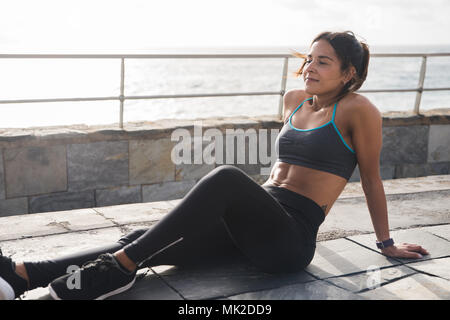 Mettre en place une jeune femme prenant une pause exercice assis dehors dans l'usure et active moderne elle-même au soleil Banque D'Images