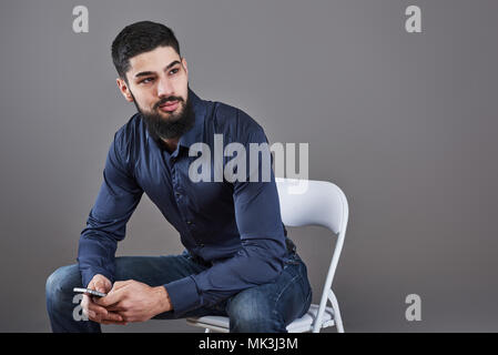 Portrait de dents beau barbu assis sur une chaise avec un téléphone sur les mains Banque D'Images