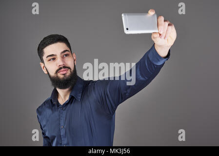Jeune hipster barbu business man photo selfies avec smart phone smiling and looking at phone contre fond studio gris Banque D'Images