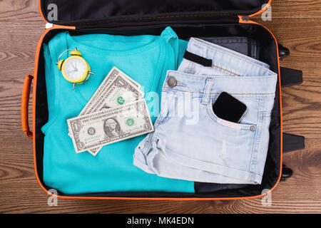 Vêtements femmes d'été et accessoires pour les voyages. Vêtements femme pliée avec réveil et de l'argent dans la valise. Haut de la vue, télévision lay. Banque D'Images