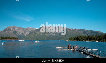 Quai sur le lac d'eau bleue avec des bateaux et forêt de chaque côté du lac de hautes montagnes rocheuses au-delà sous ciel bleu. Banque D'Images