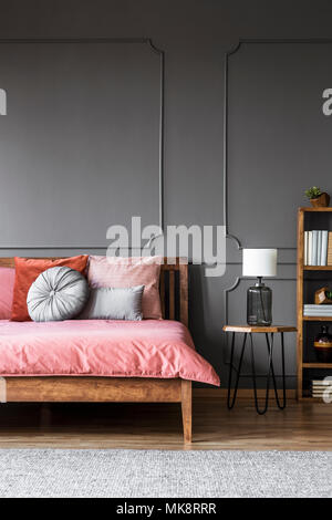 Lampe blanche sur un tabouret à côté d'un lit en bois avec draps rose foncé gris à l'intérieur chambre à coucher au moulage sur le mur Banque D'Images