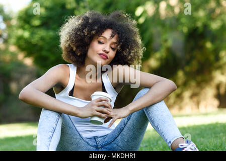 Belle jeune femme afro-américaine avec la coiffure afro. Rêve de fille aux yeux clos avec un verre à emporter dans la région de park, sitting on grass wearing ca Banque D'Images