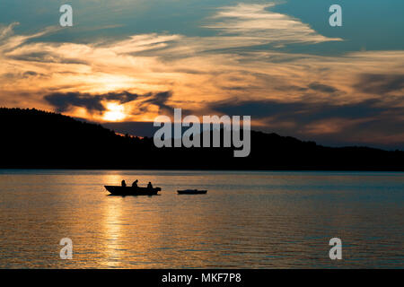 Coucher de soleil à travers les nuages, les poissons pendant que papa avec les enfants (en silhouette) à partir d'un bateau sur le lac, tout en tractant un kayak. Banque D'Images