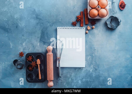 Recette de pâtisserie concept avec outils de cuisson, les ingrédients et un bloc-notes avec des pages blanches sur un fond moderne avec copie espace. Scoops, fouets, emporte-pièce, muffin tin, les oeufs et la cannelle à partir de ci-dessus. Banque D'Images