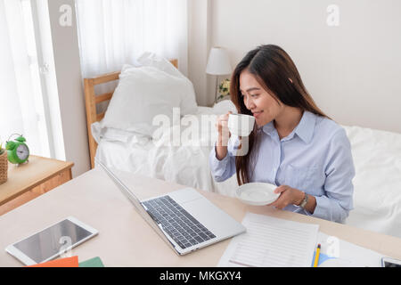 Asian businesswoman drinking coffee et tout en travaillant avec un ordinateur portable sur le tableau dans la chambre à la maison.Travail à domicile travail à domicile.concept concept Banque D'Images
