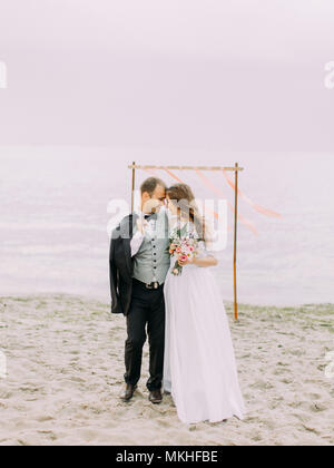 La vue sur toute la longueur de l'heureux nouveaux mariés article de tête à tête à l'avant de l'arche de mariage sur la plage. Banque D'Images