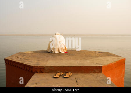 Un saint homme vêtu de blanc est en priant et en méditant sur l'un des nombreux Ghats de Varanasi en face de la rivière sacrée, le Gange en Inde. Banque D'Images