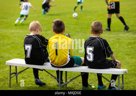 L'équipe de football pour les enfants. Les jeunes joueurs de soccer assis dans une rangée sur un banc en bois. Les garçons en noir et jaune Maillot football shirts avec des nombres. Banque D'Images