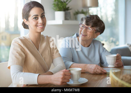 Smiling caregiver boire du thé tout en prenant soin d'une bonne femme âgée de l'hospice Banque D'Images