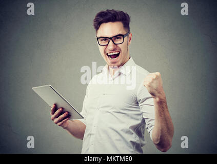 Cheerful handsome man holding pad et à la hâte avec la victoire sur fond gris. Banque D'Images