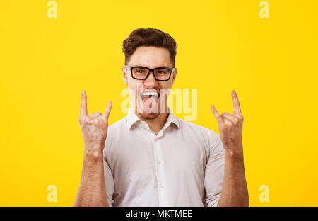 Jeune homme cool lunettes en montrant la langue et rock and roll geste sur fond jaune. Banque D'Images