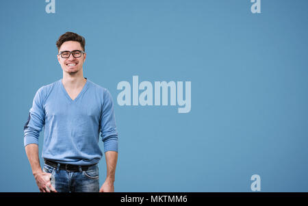 Jeune homme séduisant en chandail et lunettes à la caméra en se tenant debout sur fond bleu. Banque D'Images