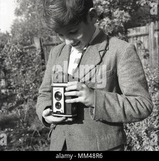 Années 1950, historiques, blazer écolier debout à l'extérieur dans un jardin à l'aide d'un appareil photo Kodak Brownie Reflex, Londres, Angleterre, Royaume-Uni. Faite au Royaume-Uni de 1946 à 1960, ce film avait un corps en bakélite de l'appareil photo et s'est tenue au niveau de la taille et comme vu un regarda le viseur. Banque D'Images