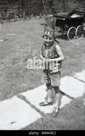 Années 1950, historiques, jeune garçon en short et portaient des sandales en extérieur dans un jardin habillé comme un indien avec un gilet et plumes hat, en Angleterre. UK. La PRAM de l'enfant est assis sur la pelouse à l'arrière-plan. Banque D'Images