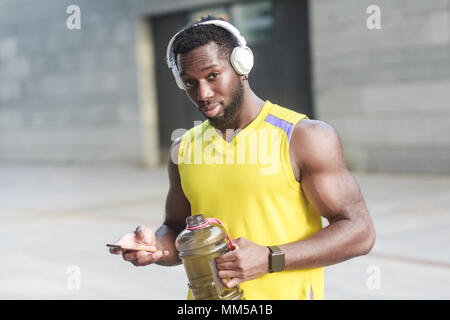 Strong homme africain de l'eau potable après l'entraînement intensif. En regardant la caméra. Holding smartphone, écouter de la musique Banque D'Images