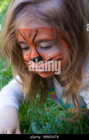 Petite fille avec un tigre dessiné sur son visage assis dans l'herbe. Le tatouage pour un jeune enfant. La créativité des enfants. Belle figure sur le visage de bébé Banque D'Images