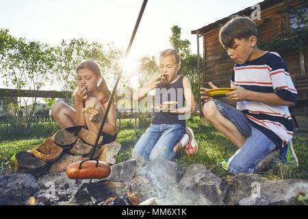 Les enfants bénéficient de camp. Frères et sœurs (famille) faire griller les saucisses sur le jardin au coucher du soleil. Banque D'Images