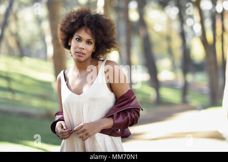 Jeune femme noire avec afro hairstyle debout dans un parc urbain. Woman wearing red mixte cuir veste et robe blanche avec fond de la ville. Banque D'Images