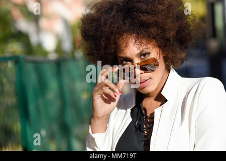 Jeune femme noire avec afro hairstyle debout dans l'arrière-plan urbain avec des lunettes de soleil aviateur. Girl wearing mixte veste blanche et noire dress posing nea Banque D'Images