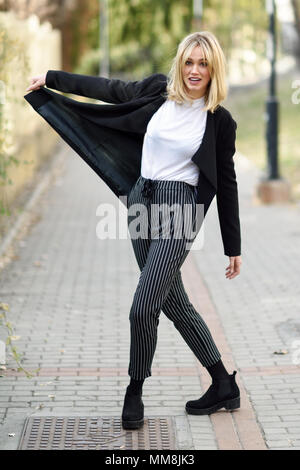 Funny blonde woman smiling en contexte urbain. Jeune fille vêtu de noir veste blazer et pantalon à rayures debout dans la rue. Jolie femelle avec Banque D'Images