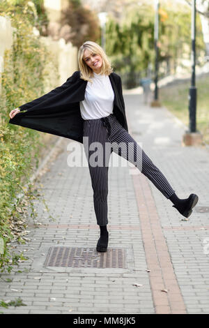 Funny blonde woman smiling en contexte urbain. Jeune fille vêtu de noir veste blazer et pantalon à rayures debout dans la rue. Jolie femelle avec Banque D'Images