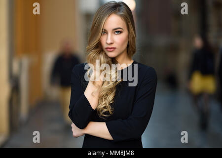 Femme blonde en contexte urbain. Belle jeune fille vêtu de noir élégante robe debout dans la rue. Jolie femme russe avec la longue chevelure ondulée h Banque D'Images