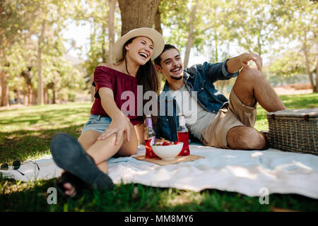 Cheerful young couple sitting in a park et faire un pique-nique. Smiling couple relaxing together avec corbeille de fruits et des bières sur couverture de pique-nique. Banque D'Images