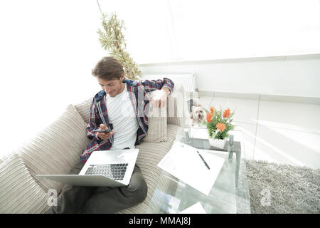 Jeune homme avec portable et smartphone assis sur un canapé dans la salle de séjour. Banque D'Images