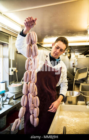 Homme portant un tablier, debout dans une boucherie, tenue grand nombre de crochets de saucisses. Banque D'Images